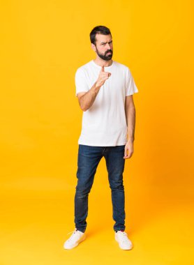 İzole edilmiş sarı arka planda sakallı bir adamın uzun pozu hayal kırıklığına uğramış ve ön tarafı işaret ediyor.
