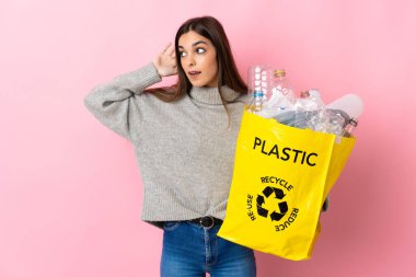 Pembe arka planda izole edilmiş plastik şişelerle dolu bir çanta taşıyan beyaz bir kadın kulağına elini sürerek bir şey dinliyor.