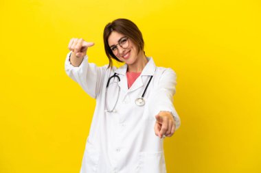 Doktor kadın sarı arka planda izole edilmiş, sana gülümserken parmak işareti yapıyor.