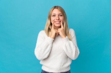 Mavi arka planda izole edilmiş genç sarışın kadın mutlu ve hoş bir ifadeyle gülümsüyor.