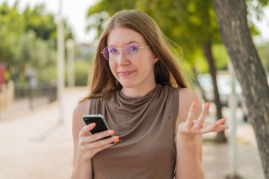 Gözlüklü Fransız kız dışarıda cep telefonu kullanıyor. Omuzlarını kaldırırken şüpheli hareketler yapıyor.