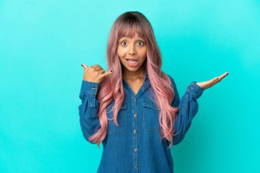 Mavi arka planda izole edilmiş pembe saçlı melez bir kadın telefon jesti yapıyor ve kuşkulu.