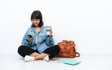 Genç bir kız öğrenci beyaz arka planda tek başına oturuyor. Elinde kahve ve cep telefonu var.