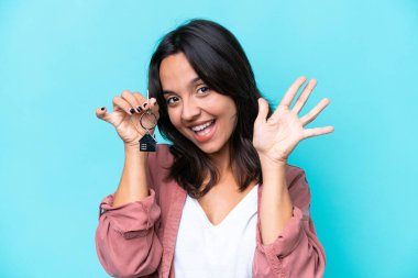 Mavi arka planda ev anahtarlarını tutan genç İspanyol kadın parmaklarıyla beşi sayıyor.