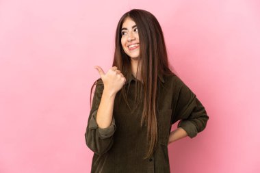 Pembe arka planda izole edilmiş genç beyaz kadın bir ürünü sunmak için kenarı işaret ediyor.