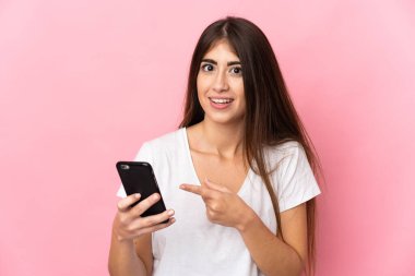 Young caucasian woman isolated on pink background using mobile phone and pointing it