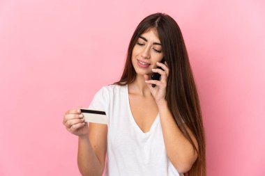 Pembe arka planda izole edilmiş genç beyaz bir kadın kredi kartıyla cep telefonuyla alışveriş yapıyor.