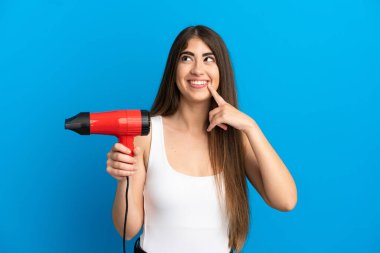 Kafkasyalı genç bir kadın elinde saç kurutma makinesiyle mavi arka planda izole edilmiş bir şekilde yukarı bakarken bir fikir düşünüyor.