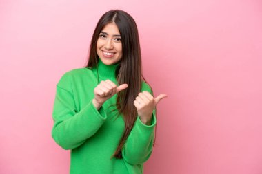 Pembe arka planda izole edilmiş genç beyaz kadın bir ürünü sunmak için kenarı işaret ediyor.