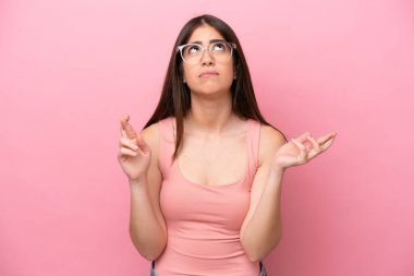 Pembe arka planda izole edilmiş genç beyaz kadın parmaklarını çapraz yapıp en iyisini diliyor.