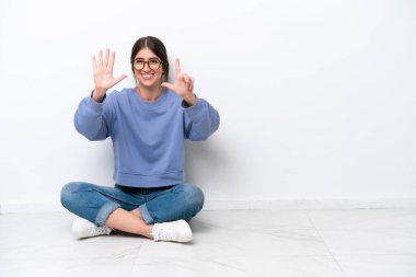 Beyaz arka planda, yerde oturan beyaz tenli genç kadın yediyi parmaklarıyla sayıyor.