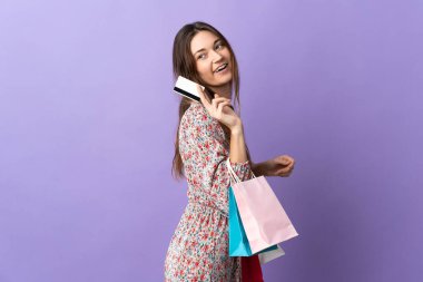 Mor arka planda izole edilmiş genç bir İrlandalı kadın elinde alışveriş çantaları ve bir kredi kartı tutuyor.