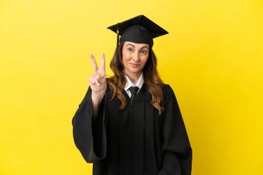 Sarı arka planda izole edilmiş orta yaşlı üniversite mezunu. Gülümsüyor ve zafer işareti gösteriyor.