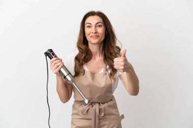 Orta yaşlı beyaz kadın el blenderi kullanıyor. Beyaz arka planda izole edilmiş. Başparmaklar havada çünkü iyi bir şey oldu.