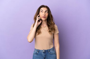 Mor arka planda izole edilmiş cep telefonu kullanan orta yaşlı beyaz kadın yukarı bakarken bir fikir düşünüyor.