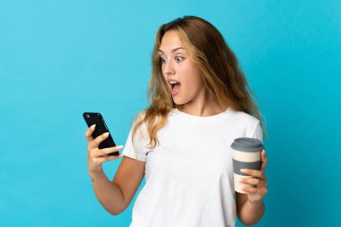Mavi arka planda izole edilmiş genç sarışın kadın elinde kahve ve bir cep telefonu tutuyor.