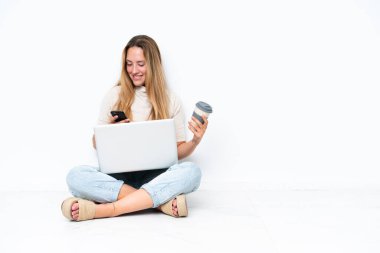 Young woman with laptop sitting on the floor isolated on white background holding coffee to take away and a mobile