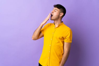 Mor arka planda izole edilmiş genç Kolombiyalı adam esniyor ve ağzını eliyle açık tutuyor.