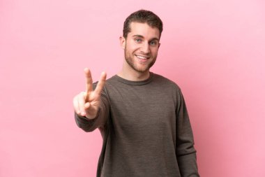 Pembe arka planda izole edilmiş genç beyaz adam gülümsüyor ve zafer işareti gösteriyor.