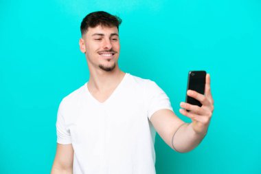 Kafkasyalı yakışıklı adam mavi arka planda izole edilmiş cep telefonuyla selfie çekiyor.