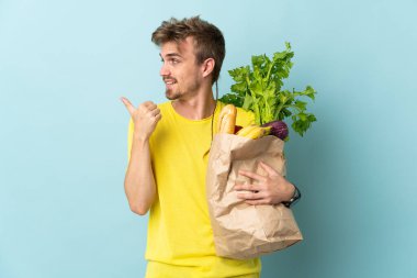 Sarışın bir adam mavi arka planda izole edilmiş bir paket yiyecek alıyor. Yan tarafı göstererek ürünü sunuyor.