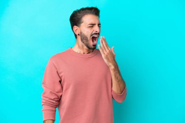 Kafkasyalı genç adam mavi arka planda izole edilmiş esniyor ve geniş açık ağzını eliyle kapatıyor.