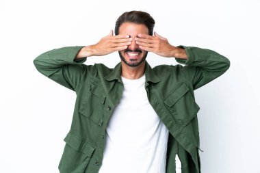 Beyaz arka planda izole edilmiş beyaz tenli genç bir adam. Gözleri elleriyle kaplıyor ve gülümsüyor.