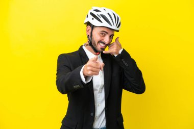 Bisiklet kasklı beyaz bir adam sarı arka planda izole edilmiş telefon hareketi yapıyor ve ön tarafı işaret ediyor.