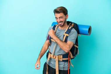 Mavi arka planda büyük bir sırt çantası olan genç dağcı zaferi kutluyor.