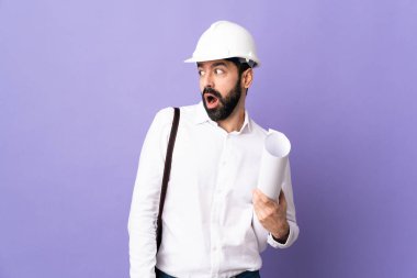 Miğferli genç bir mimar. Mor arka plan üzerinde planlar tutuyor. Yan tarafa bakarken sürpriz jest yapıyor.