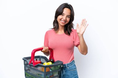 Genç bir kadın elinde yiyecek dolu bir sepet tutarken mutlu bir ifadeyle selamlıyor.