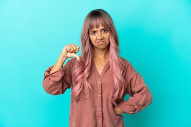 Mavi arka planda izole edilmiş pembe saçlı, melez genç bir kadın olumsuz bir ifadeyle baş parmağını aşağı gösteriyor.