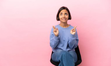 Genç melez kadın pembe arka planda izole edilmiş bir sandalyede oturuyor ve en iyisini diliyor.