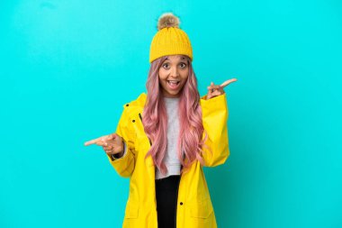 Pembe saçlı, yağmura dayanıklı bir ceket giyen mavi arka planda izole edilmiş genç bir kadın parmak uçlarını işaret ediyor ve mutlu.