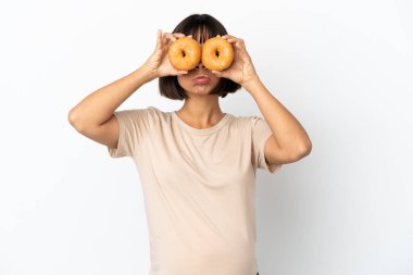 Genç melez hamile kadın, beyaz arka planda izole edilmiş üzgün bir ifadeyle gözlerinde donut tutuyordu.