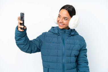 Beyaz arka planda izole edilmiş, kış kulaklığı takan genç İspanyol kadın selfie çekiyor.
