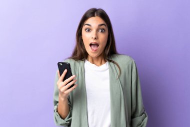 Mor arka planda izole edilmiş, şaşırmış ve şok olmuş bir yüz ifadesiyle cep telefonu kullanan beyaz bir kadın.