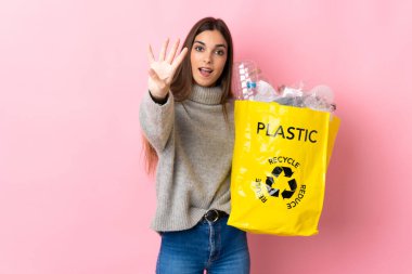 Pembe arka planda izole edilmiş plastik şişelerle dolu bir çanta taşıyan beyaz bir kadın mutlu ve parmaklarıyla dört tane sayıyor.