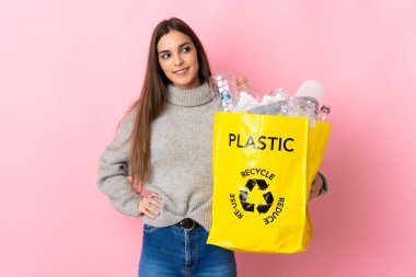Elinde plastik şişelerle dolu bir çanta tutan beyaz bir kadın pembe arka planda izole edilmiş, kolları kalçasında poz veriyor ve gülümsüyor.