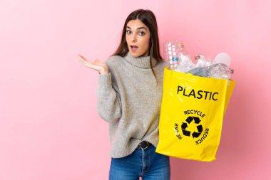 Elinde plastik şişelerle dolu bir çanta tutan beyaz bir kadın. Pembe arka planda izole edilmiş, şok olmuş bir yüz ifadesi var.