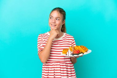 Mavi arka planda yalıtılmış waffle tutan küçük beyaz kız yana bakıyor ve gülümsüyor.