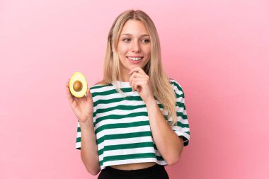 Pembe arka planda izole edilmiş bir avokado tutan genç beyaz kadın yana bakıyor ve gülümsüyor.