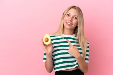 Elinde pembe arka planda izole edilmiş avokado tutan beyaz bir kadın bir ürünü sunmak için kenarı işaret ediyor.