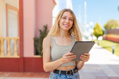 Açık havada elinde mutlu bir ifadeyle tablet tutan genç sarışın kadın.
