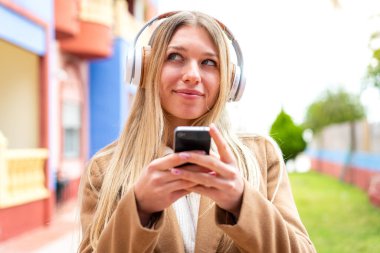 Genç, güzel, sarışın bir kadın açık havada müzik dinliyor ve düşünüyor.