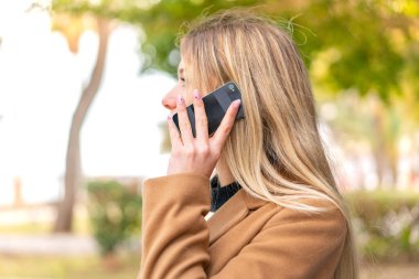 Açık havada genç, güzel sarışın bir kadın cep telefonuyla biriyle sohbet ediyor.