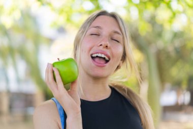 Açık havada elinde mutlu bir ifadeyle bir elma tutan genç sarışın kadın.