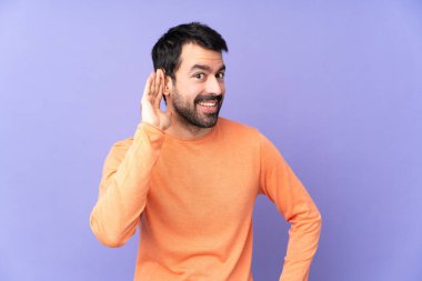 Mor arka planda beyaz tenli yakışıklı bir adam elini kulağına koyarak bir şeyler dinliyor.