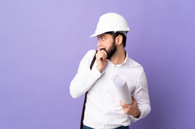 Miğferli genç bir mimar. Mor arka planda izole edilmiş planları var. Ağzı örtüyor ve yan tarafa bakıyor.