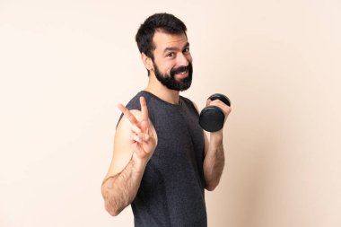 Beyaz sakallı sporcu arka planda halter kaldırıyor. Gülümsüyor ve zafer işareti gösteriyor.